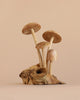 The Small Wooden Natural Mushroom Trio features three wooden mushrooms with light brown caps and stems, artfully carved from a single piece of wood and arranged on a textured driftwood base, set against a plain beige background.