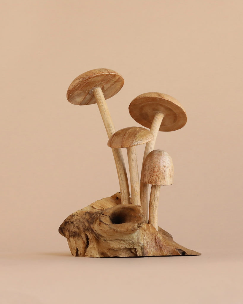 The Small Wooden Natural Mushroom Trio features three wooden mushrooms with light brown caps and stems, artfully carved from a single piece of wood and arranged on a textured driftwood base, set against a plain beige background.