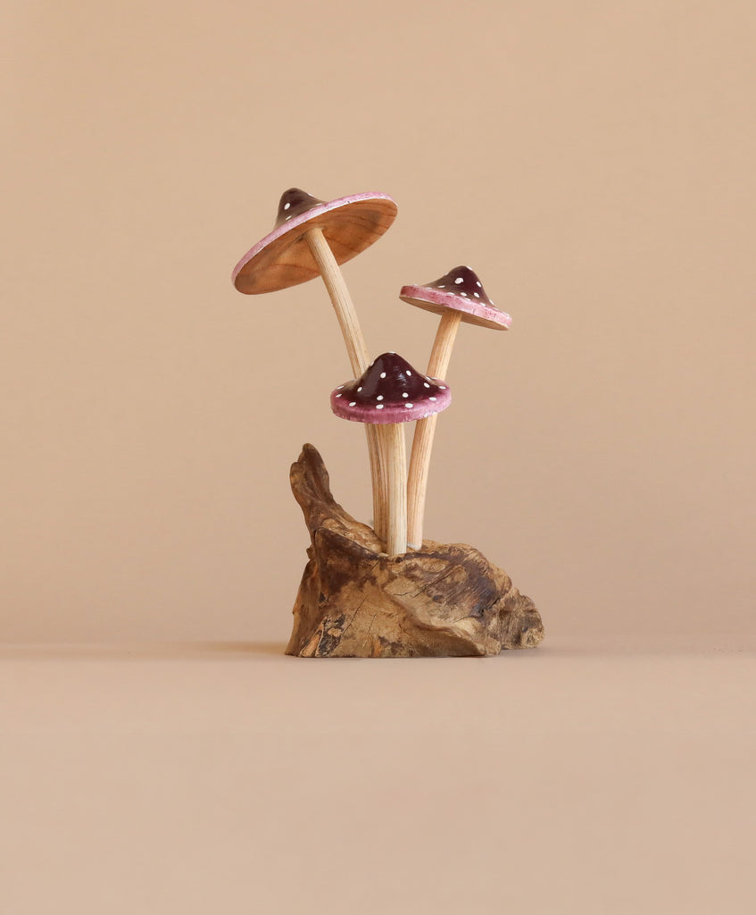 The Small Wooden Purple Mushroom Trio features three handcrafted wooden mushrooms, each with brown stems and dark purple caps edged in pink, growing from a piece of brown wood. This decorative figurine rests on a driftwood base, set against a light beige background for a minimalist and natural look.