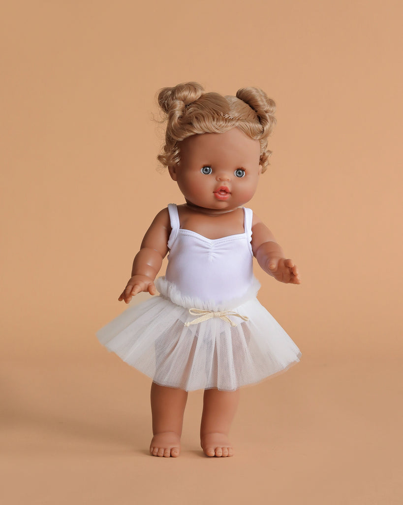 The Minikane Standing Doll (14") – Louisa with Clothing features brown hair, blue eyes, and a white ballerina outfit with tulle skirt. This barefoot, anatomically correct doll is beautifully made in Spain and poses against a beige background.
