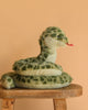 The Slyde Spotted Snake Plush Toy, featuring green spots and a red tongue, sits coiled on a wooden stool against a peach backdrop—a charming addition to any plush collection.