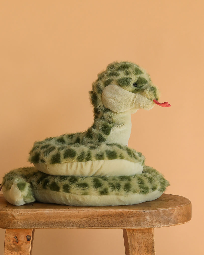 The Slyde Spotted Snake Plush Toy, featuring green spots and a red tongue, sits coiled on a wooden stool against a peach backdrop—a charming addition to any plush collection.