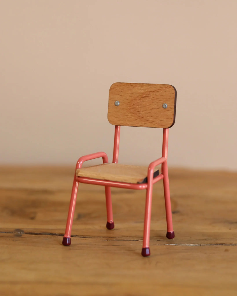 A small wooden chair with a light brown seat and pink metal legs stands on a wooden surface next to the Maileg Bathroom Set, set against a beige background.