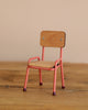 The Maileg Mouse Size School Chair - Coral, with its pink metal legs and matching frame, rests on a wooden surface. Featuring a simple design with a rectangular backrest and flat seat, this charming piece could easily complement a table and chair set against the plain beige backdrop.