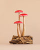 The Small Wooden Red Mushroom Trio features three red and white spotted mushrooms with tall, slender stems arranged on a driftwood base, set against a plain beige background for a charming decorative display.