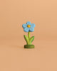 The Bumbu Handmade Wooden Flower Festive Forget-Me-Not features blue petals, a yellow center, green leaves, and a green base—perfect for Montessori, Waldorf play, or celebration rings. Stands upright against a peach-colored background.