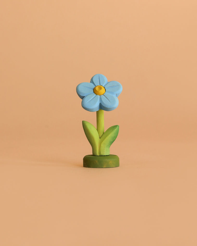 The Bumbu Handmade Wooden Flower Festive Forget-Me-Not features blue petals, a yellow center, green leaves, and a green base—perfect for Montessori, Waldorf play, or celebration rings. Stands upright against a peach-colored background.