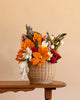 The Basket of Felt Flowers, a woven basket brimming with vibrant orange, white, red, and purple wool felt blooms, rests on a wooden table set against a plain beige background. This everlasting bouquet brings a charming and colorful accent to any space.