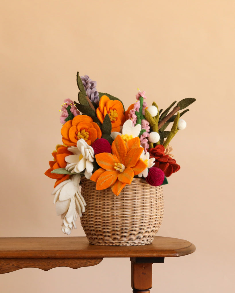 The Basket of Felt Flowers, a woven basket brimming with vibrant orange, white, red, and purple wool felt blooms, rests on a wooden table set against a plain beige background. This everlasting bouquet brings a charming and colorful accent to any space.