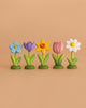 The Bumbu Handmade Wooden Celebration Ring features five upright wooden flowers in blue, purple, yellow, pink, and white on green bases—eco-friendly toys that inspire creative Montessori play against a simple beige background.