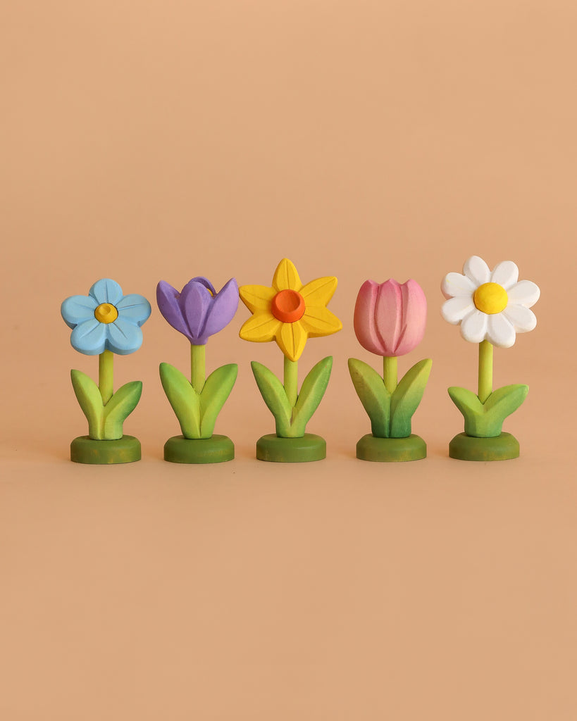 The Bumbu Handmade Wooden Festive Flower Set displays five eco-friendly wooden flowers—blue, purple, yellow, pink, and white—with unique petal shapes and green stems and bases arranged in a row on a light brown background.