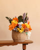 The Basket of Felt Flowers, featuring a woven basket overflowing with vibrant wool felt blooms in shades of orange, yellow, white, purple, and green, sits on a wooden table against a light beige background—creating an everlasting bouquet that brightens any space.