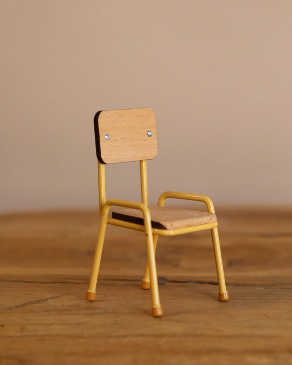 The Maileg Mouse Size School Chair - Yellow is a charming wooden toy with hand-painted details, featuring yellow metal legs and backrest, and sits quaintly on a wooden surface against a neutral beige background.