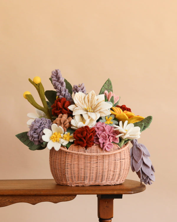 The Basket of Felt Flowers - Final Sale, featuring a vibrant arrangement of felt flowers and green felt leaves in a woven basket, sits on a wooden table against a plain light brown background, creating a charming eco-friendly floral display.