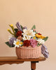 The Basket of Felt Flowers - Final Sale, featuring a vibrant arrangement of felt flowers and green felt leaves in a woven basket, sits on a wooden table against a plain light brown background, creating a charming eco-friendly floral display.