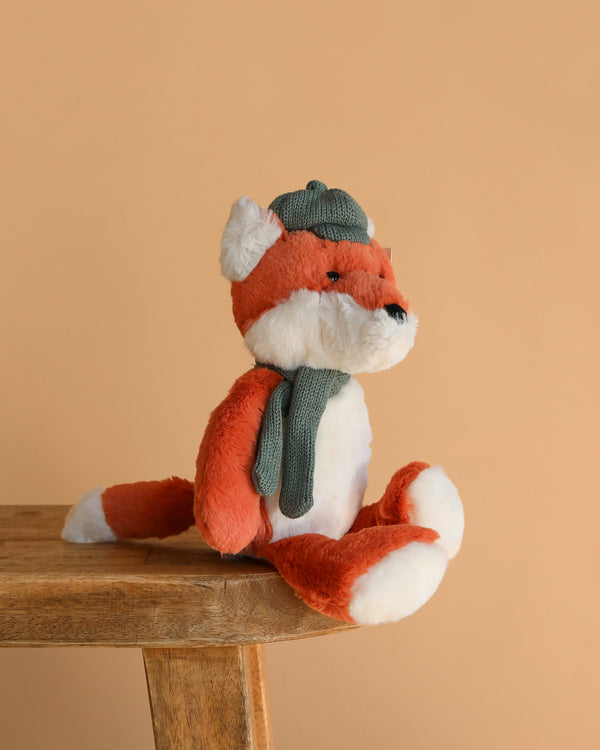 The Frederick Fox Plush Toy, featuring orange and white fur and dressed in a green knit hat and scarf, sits on a wooden table against a plain beige background.