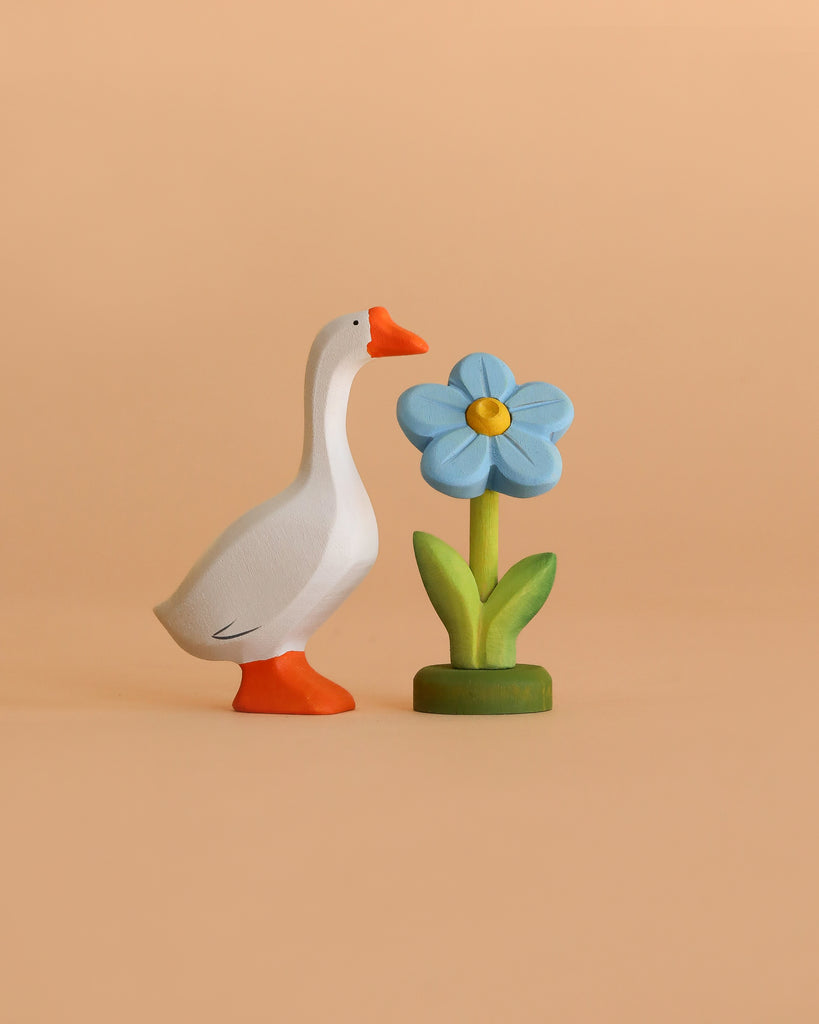 The Bumbu Handmade Wooden Spring Set features a hand-carved white goose with orange beak and feet beside a blue flower with green leaves, all set against a beige background—perfect for Montessori toy collections.