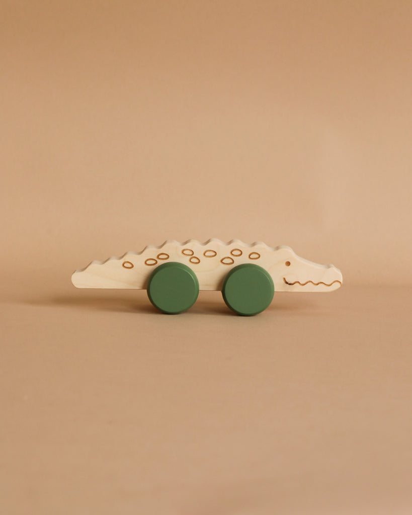 A minimalist, handmade in Poland, Wooden Crocodile Push Toy featuring two green wheels. With simple, etched details on its body to represent the crocodile's features, this push toy is perfect for developing fine motor skills and is set against a neutral-colored background.