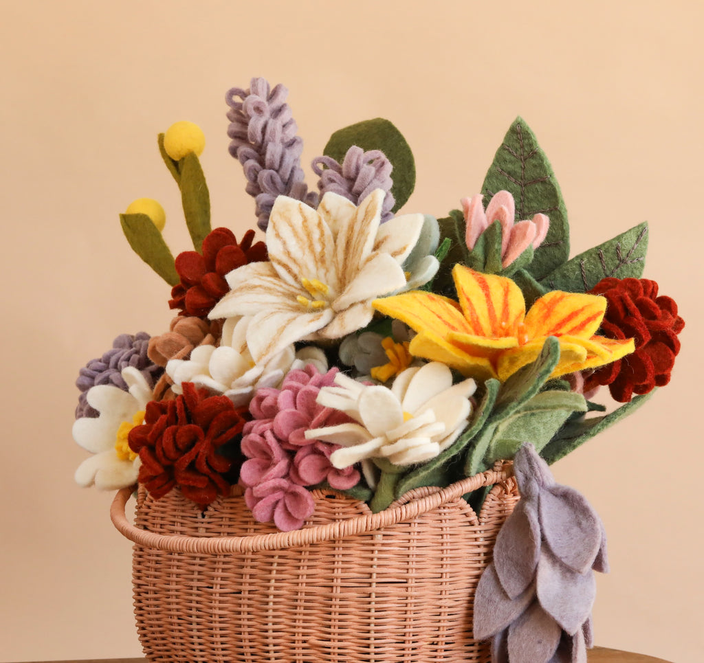 Introducing our Basket of Felt Flowers - Final Sale: a woven basket brimming with colorful felt flowers—yellow, white, purple, red, and pink blooms enhanced by green felt leaves—all arranged against a beige backdrop. This eco-friendly display instantly brightens any space and adds cheerful charm.