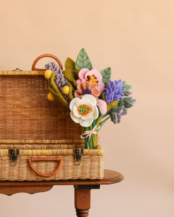 A Handcrafted Felt Flower Bouquet - Final Sale sits in a wicker basket on a wooden table, displaying vibrant blooms in shades of purple, yellow, pink, and white with green felt leaves, set against a plain beige background.