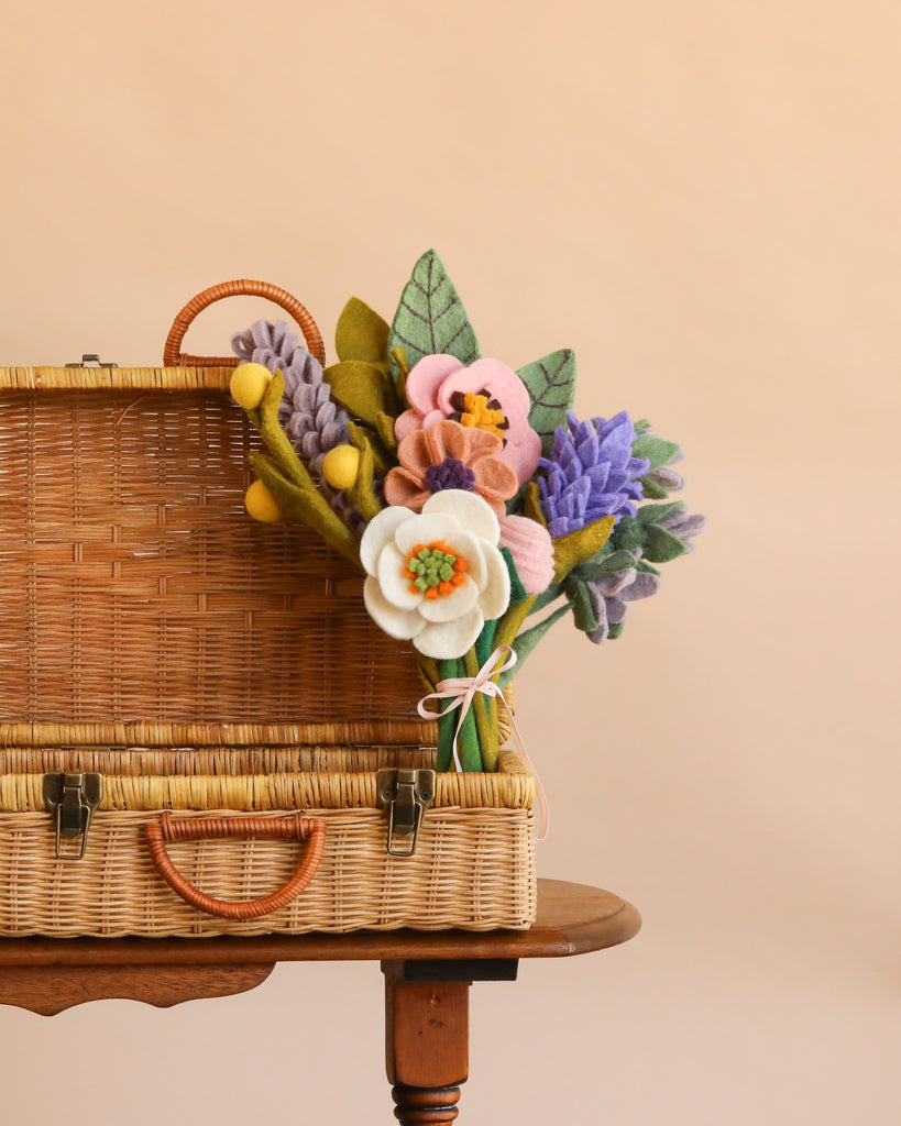 A Handcrafted Felt Flower Bouquet - Final Sale sits in a wicker basket on a wooden table, displaying vibrant blooms in shades of purple, yellow, pink, and white with green felt leaves, set against a plain beige background.