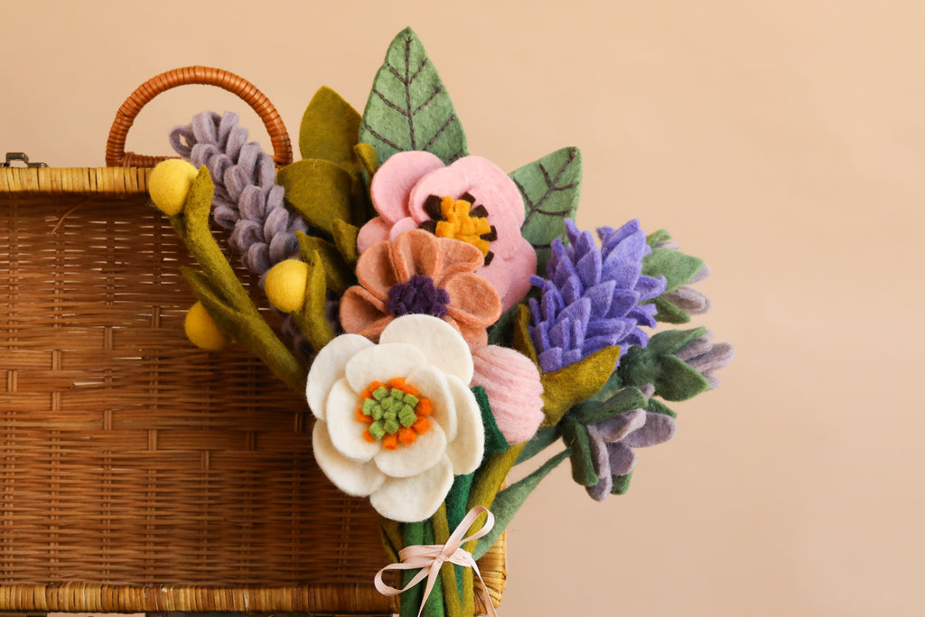 The Handcrafted Felt Flower Bouquet - Final Sale showcases a vibrant mix of white, pink, purple, and yellow felt blooms with green leaves, artfully arranged and tied with a pale ribbon. This cheerful bouquet is styled against a wicker basket and beige background.