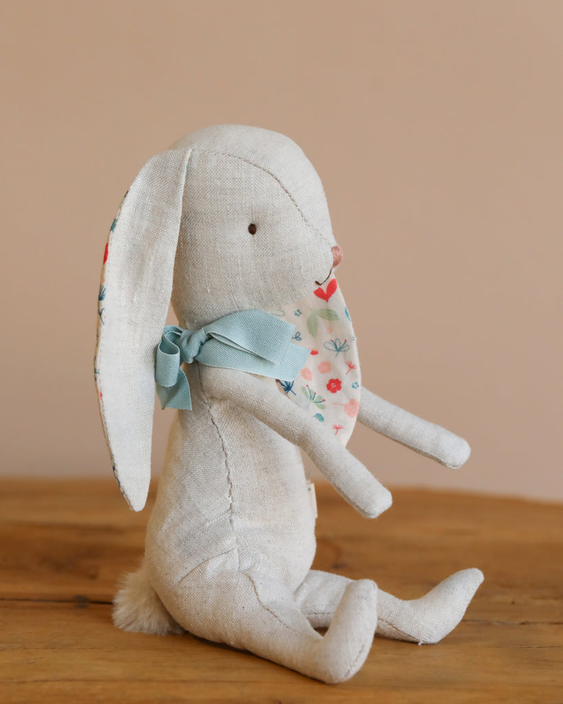 A handmade Maileg Bunny Albin, crafted from soft linen, sits on wood. It features long floppy ears with floral inner patterns and a blue bow around its neck against a plain beige wall.