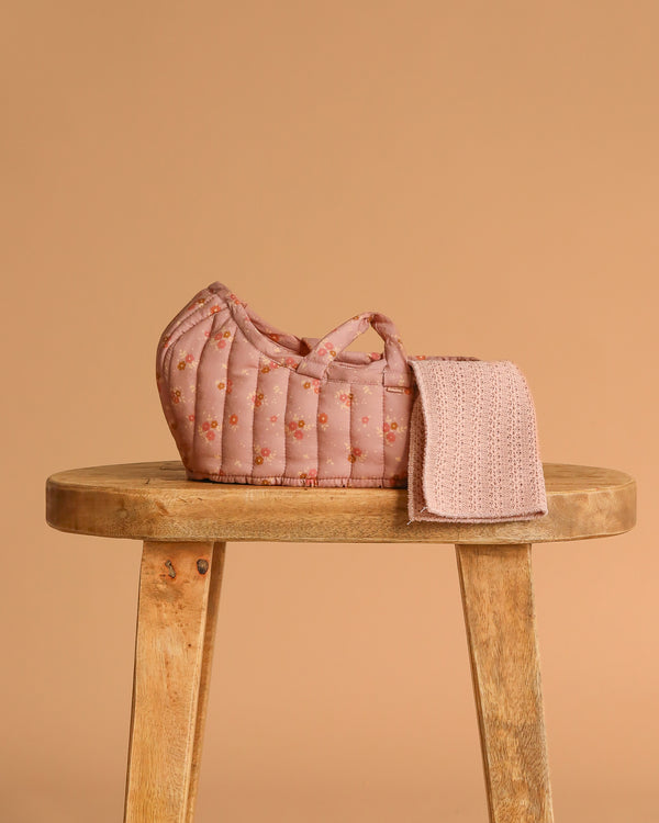 The Maileg Carry Cot, Large - Rose, a quilted pink floral basket with handles, sits on a wooden stool with a textured pink cloth draped over the side against a plain warm beige background—ideal as a portable doll bed.