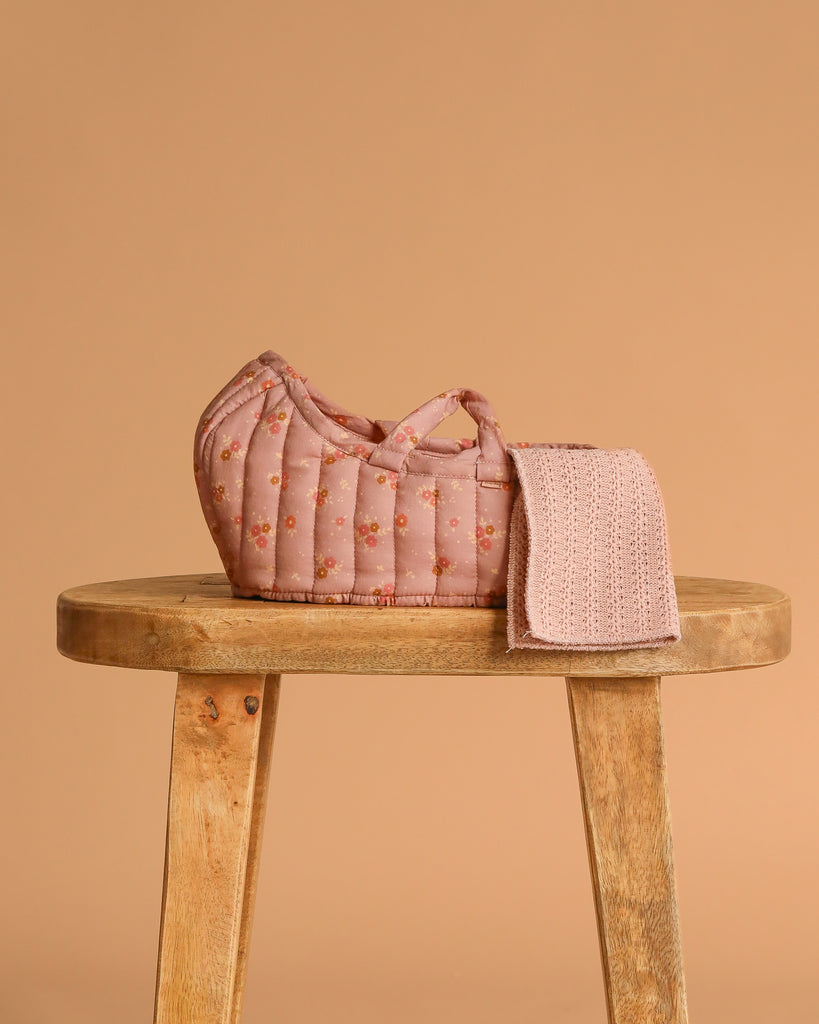 The Maileg Sleeping Kitten in Carry Cot, featuring a small quilted pink basket with floral patterns and a textured pink cloth, rests on a wooden stool against a plain beige background.