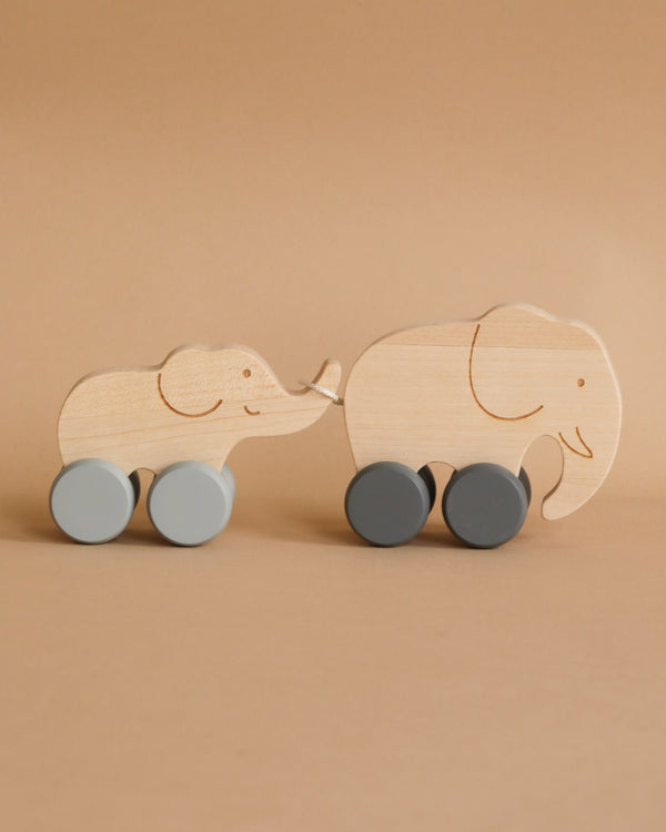 Two wooden Mom and Baby Elephant Push Toy Set with wheels are set against a beige background. The smaller elephant, with light blue wheels, holds the tail of the larger one, equipped with dark gray wheels. Engraved details on the wood depict their ears, eyes, and trunks. These toys are safe for children and help develop fine motor skills.