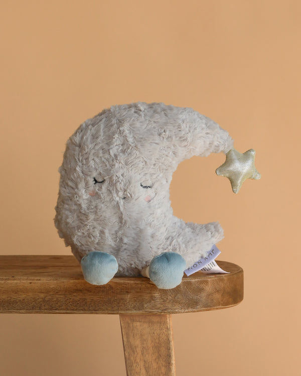 The Moon Beam Plush Toy, featuring closed eyes, blushing cheeks, blue feet, and a shiny star at its tip, sits on a wooden stool—a perfect celestial snuggle buddy against a beige background.