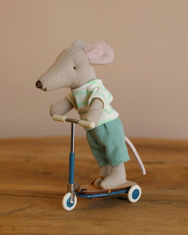 A joyful stuffed mouse in a green striped shirt and blue pants rides the Maileg Mouse Size Scooter / Kickboard - Blue, radiating a playful look on a detailed wooden surface.