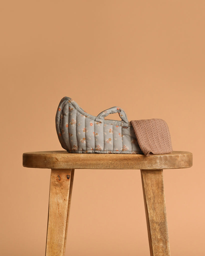 A Maileg Sleeping Kitten in Carry Cot, featuring a small quilted gray bed with a floral pattern and a brown knitted blanket, sits on a wooden stool against a solid beige background.