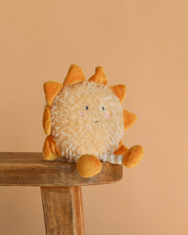The Sun Beam Plush Toy, with a smiling face, orange rays, and soft yellow arms and legs, sits on a wooden bench against a light orange background—perfect as cheerful nursery decor.