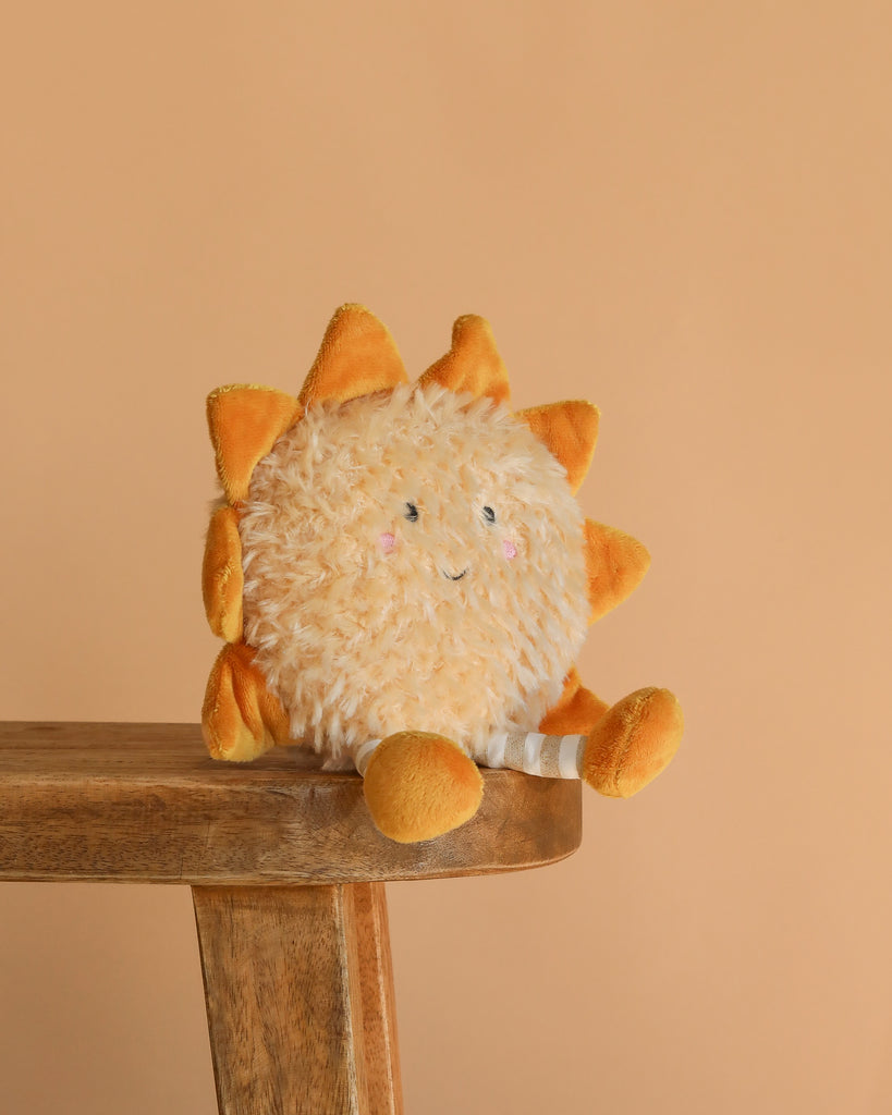 The Sun Beam Plush Toy, with a smiling face, orange rays, and soft yellow arms and legs, sits on a wooden bench against a light orange background—perfect as cheerful nursery decor.