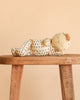 The Olli Ella Dozy Dinkum – Custard Sweetheart, dressed in patterned pajamas, lies on its side on a wooden stool against a peach-colored background.