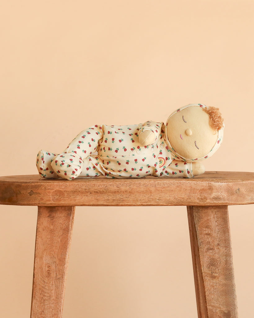 The Olli Ella Dozy Dinkum – Custard Sweetheart, dressed in patterned pajamas, lies on its side on a wooden stool against a peach-colored background.