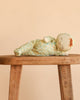 The Olli Ella Dozy Dinkum - Pickle Blossom, dressed in a charming yellow floral outfit with a matching bonnet, lies on its side atop a rustic wooden stool against a beige background.