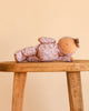 The Olli Ella Dozy Dinkum - Tootsie Briar doll, dressed in a purple floral outfit, lies on its side on a wooden stool against a beige background.
