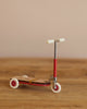 The Maileg Mouse Size Scooter / Kickboard - Red, with white wheels and wooden details, stands on a wooden surface. It features a sleek metal stick with small deck and handlebars, set against a plain beige wall.