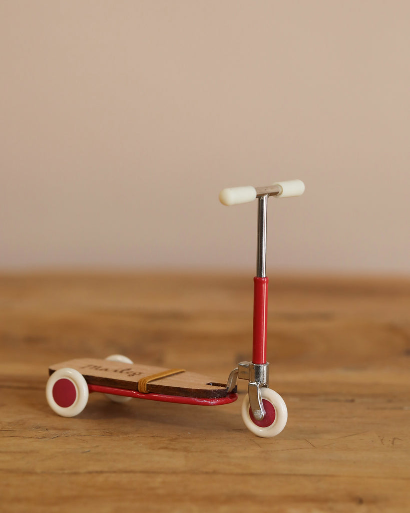 The Maileg Mouse Size Scooter / Kickboard - Red, with white wheels and wooden details, stands on a wooden surface. It features a sleek metal stick with small deck and handlebars, set against a plain beige wall.