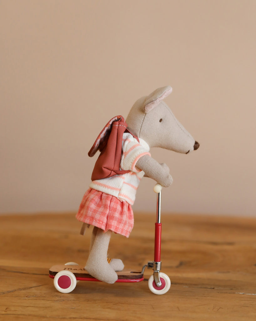 A stuffed animal resembling a mouse rides a Maileg Mouse Size Scooter - Red, dressed in a striped shirt and plaid skirt. It carries a small brown backpack against a warm-toned background with charming wooden details.