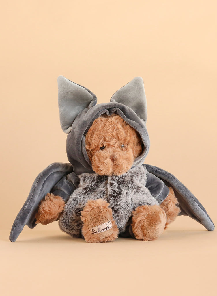 The Bukowski - Bear In Bat Costume features a plush teddy bear dressed in a gray bat outfit with large ears and wings, ideal for nighttime cuddles, and is pictured against a plain beige background.