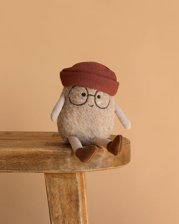 The Spud Bud Potato Plush Toy, featuring glasses, a red knit hat, and brown shoes, sits on a wooden bench against a beige background—a quirky and cuddly gift idea.