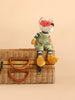 Bukowski - Foxy, a plush fox toy with stitched eyes dressed in green overalls, sits on a closed wicker suitcase with leather handles against a beige background.