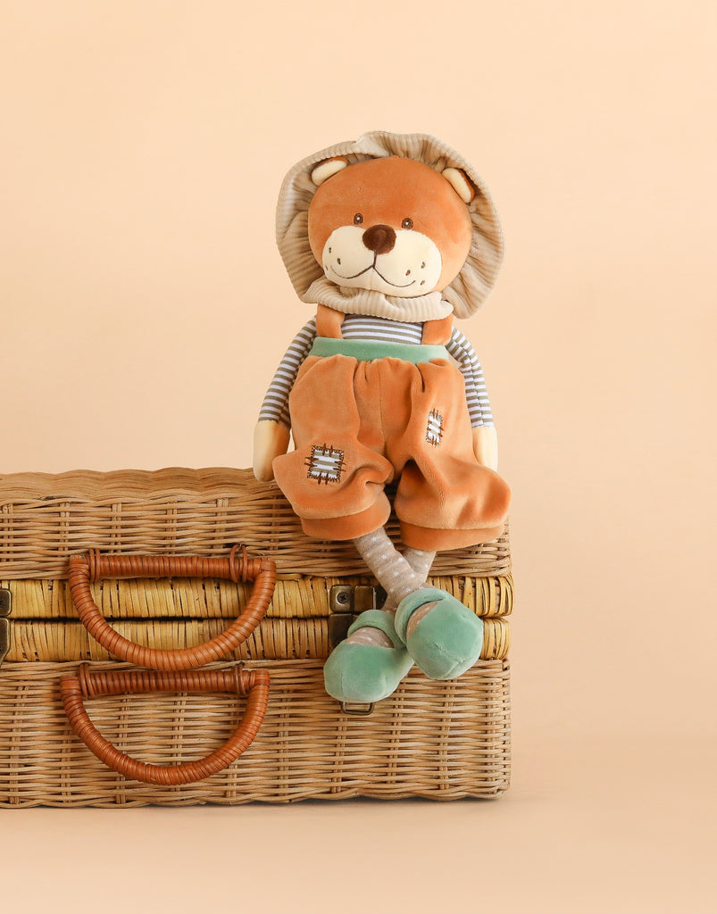 The Bukowski - Happy Mael Lion is an adorable plush stuffed toy featuring a lion in orange overalls and a striped shirt, sitting on a woven wicker suitcase with tan handles against a beige background.