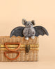 The Bukowski - Baltazar Bat, a gray plush cuddly toy with large ears and outstretched wings, sits atop a closed wicker basket against a plain beige background.