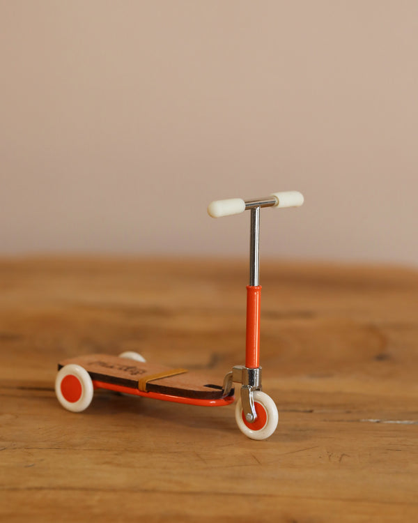 The Maileg Mouse Size Scooter in orange rests on wood, showing off its wooden details against a plain beige background. It features a T-shaped handlebar and white wheels, making it perfect for tiny riders.