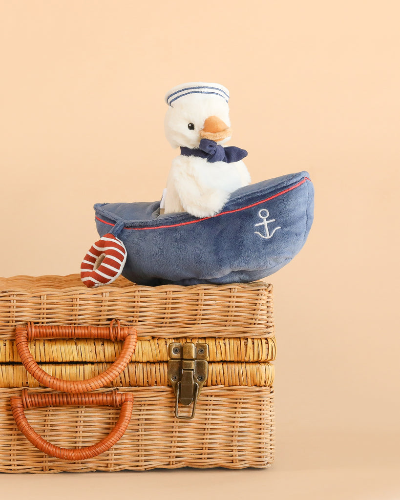 The Mon Ami - Sailor Duck, dressed in a charming sailor outfit, sits in a plush blue boat adorned with an anchor emblem, creating an appealing nautical stuffed animal display atop a wicker suitcase against a beige background.
