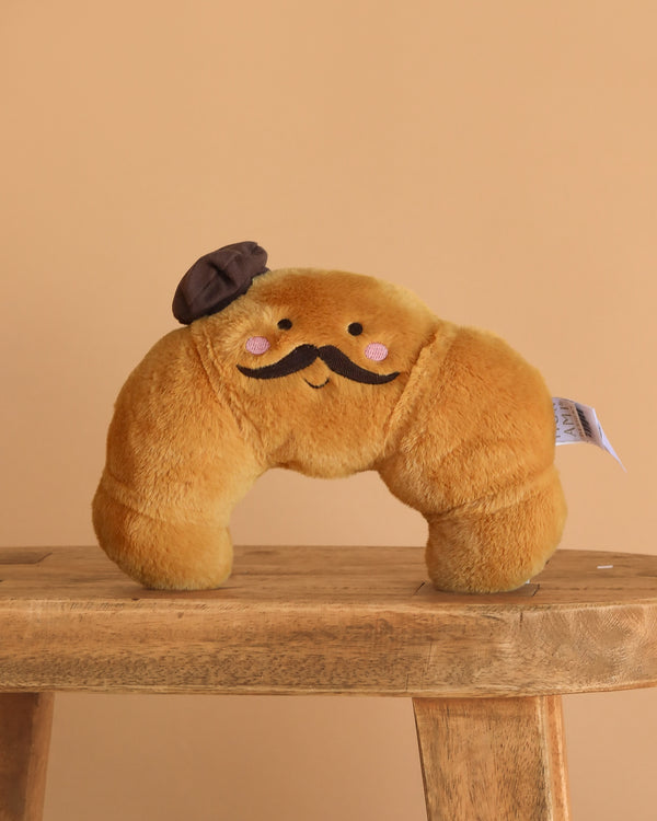 The Monsieur Croissant Plush Toy, featuring a smiling face, pink cheeks, black mustache, and brown beret, sits on a wooden stool against a plain beige background.
